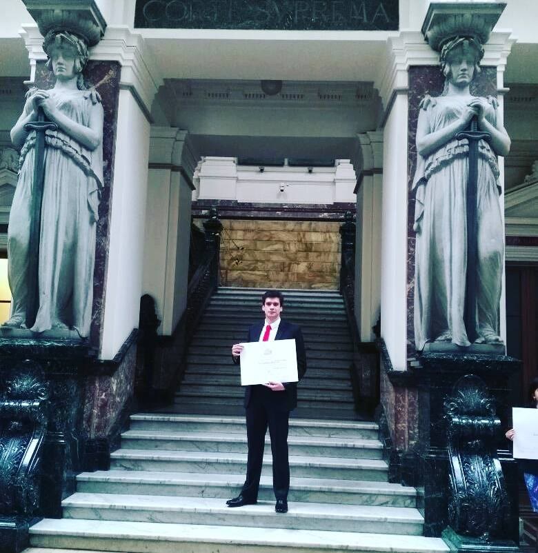 Un recién licenciado sostiene su certificado en la escalinata de mármol de la Corte Suprema, entre dos estatuas